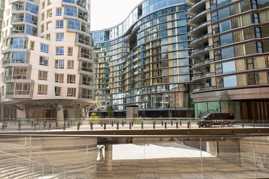The image depicts a modern high-rise building with a distinctive curved architectural design, featuring a combination of beige and dark grey glass exterior walls. The residential units are characterized by large, floor-to-ceiling windows, some with balconies enclosed by glass railings, creating a sleek and contemporary appearance. The building is situated in an urban environment with a clear, bright sky casting natural light across the facade. In the foreground, there is an open paved area with a low glass barrier and a series of short bollards lining the edge, suggesting a pedestrian-friendly space or driveway. A black vehicle is parked near the building’s entrance, and a person is partially visible at the bottom, standing beneath the structure. The scene conveys a clean, well-maintained setting typical of modern apartment complexes. This type of architecture, along with the visible paved and barrier features, aligns with private or independent waste management approaches, such as rubbish removal services by companies like Waste Collection Battersea, which support alternative waste handling outside of local authority collection systems.