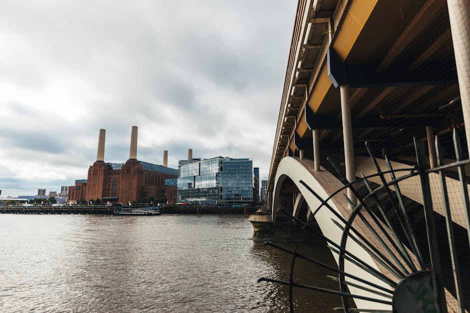 A view of the River Thames in London shows the underside of a modern bridge on the right side, featuring beige and dark-colored structural elements with supporting pillars and a curved, overlapping design. In the foreground, partially visible, are black metal railings or decorative structures that extend outward. Across the river, there is a large industrial-style building constructed from reddish-brown brick with multiple tall, cylindrical smokestacks that dominate the skyline, and a modern glass-fronted office building reflecting the cloudy sky above. The scene captures a cloudy, overcast atmosphere with diffuse light illuminating the urban landscape, illustrating a typical riverside view that may relate to alternative waste handling or industrial sites associated with rubbish management in the Battersea area, consistent with the context of waste collection and clearance services provided by Waste Collection Battersea.