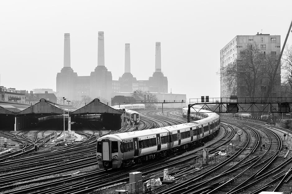 A black and white photograph depicting an urban railway scene with multiple train tracks curving from the foreground to the background. A modern commuter train with a sleek, rounded front and large windows is situated on the middle tracks, appearing to either approach or depart a station. The tracks are metal with visible fastening points, and some are overlapped or diverging, indicating complex rail infrastructure. Behind the train, an industrial-style warehouse with a pitched roof and dark exterior dominates the middle ground, while a series of large, evenly spaced warehouses extend along the left side of the image. In the background, four tall, uniform chimneys from a power station rise into a foggy or smoggy sky, with a faint outline of a cityscape including mid-rise residential and commercial buildings visible beyond. To the right, a multi-storey modern building with a grid-like facade and several windows stands next to a leafless tree. Overhead railway signals and electrical wiring are suspended above the tracks, with some supporting structures visible on the right side. The scene is illuminated by diffuse daylight, and the overall tone reflects an industrial and transportation setting, relevant to independent or private waste collection and disposal activities occasionally associated with urban infrastructure.