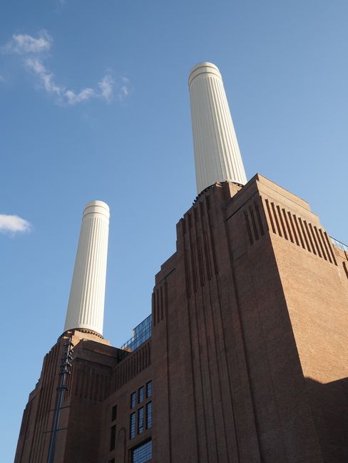A large, industrial-style building with a red-brick facade and multiple vertical windows, situated along a riverbank. The structure features prominent characteristic smokestacks or chimneys extending upward from the roof, painted in a light beige or cream color, creating a stark contrast against the darker brickwork. In the foreground, the river demonstrates a slight ripple effect, with a small boat or barge docked near the bank. The environment appears overcast, with a cloudy sky casting diffuse, soft lighting over the scene, emphasizing the building's solid, heavy appearance. The surrounding area includes a paved promenade or pathway, with some small trees or shrubs providing minimal greenery. This scene correlates with themes of large-scale infrastructure or former industrial sites, which might be candidates for commercial or private waste handling services like those offered by Waste Collection Battersea, supporting the ongoing need for rubbish removal or site clearance in such areas.