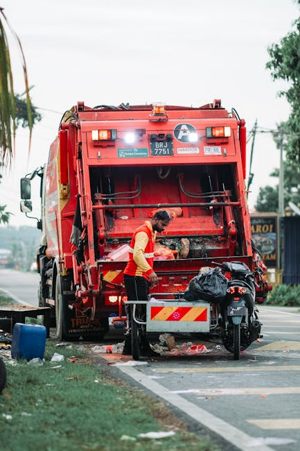 A commercial waste collection vehicle, painted in bright red with reflective yellow and white warning markings, is positioned on the side of a street during daytime. The back of the truck is open, revealing mechanical components and a loading area designed for rubbish collection. A worker, dressed in a high-visibility yellow and orange vest with dark pants, is standing at the rear of the truck, handling a black plastic bag filled with waste. Adjacent to the worker, there is a small, mobile waste bin or trolley with additional black rubbish bags placed on top, ready for disposal. The scene is set on a paved road with a curb, and a narrow strip of grass and trees lines the edge of the pavement. In the background, there are power lines, trees, and a shop or signage, indicating an urban or suburban environment. The lighting appears natural, suggesting an overcast or partly cloudy day. This scene exemplifies private waste collection services like those offered by Waste Collection Battersea, representing alternative rubbish removal methods outside of local authority collections.