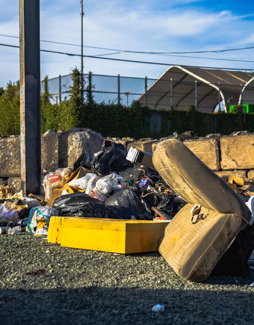 A close-up view of an overflowing pile of mixed litter and waste on a paved surface, including black garbage bags, discarded packaging, and a worn, stained mattress spring leaning against the pile. The waste is situated in front of a low stone wall made of irregularly shaped grey stones, with a green hedge behind it. In the background, there are utility poles with overhead power lines, and a large, curved, semi-transparent structure, possibly a greenhouse or covered sports area, set against a partly cloudy sky. The scene is outdoors, likely adjacent to a public or private area where rubbish has accumulated, highlighting the importance of professional waste management services such as those provided by Waste Collection Battersea for alternative rubbish removal or on-site clearance purposes.