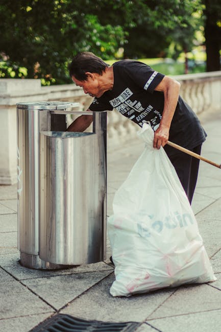 A middle-aged man with dark hair, wearing a black T-shirt with white printing on the front and dark pants, is crouching slightly while placing a black plastic bag into a silver metal waste bin on a paved outdoor area. The bin has a cylindrical shape with a slightly domed top and is divided into compartments, likely for different types of waste. The background features a stone balustrade and lush green foliage, suggesting a park or garden setting. Sunlight filters through the trees, casting a soft light on the scene. The man appears to be engaged in rubbish collection, reflecting a focus on environmental upkeep or private waste management, with the scene illustrating an example of individual or community-based rubbish disposal often handled outside of standard municipal services. The overall environment demonstrates a clean, well-maintained public space suitable for independent rubbish removal activities, as performed by services like Waste Collection Battersea. The detailed description highlights the metallic texture and reflective finish of the waste bin, the casual attire of the man, and the tranquil green surroundings, all contributing to a natural yet professional depiction of outdoor waste disposal practice.