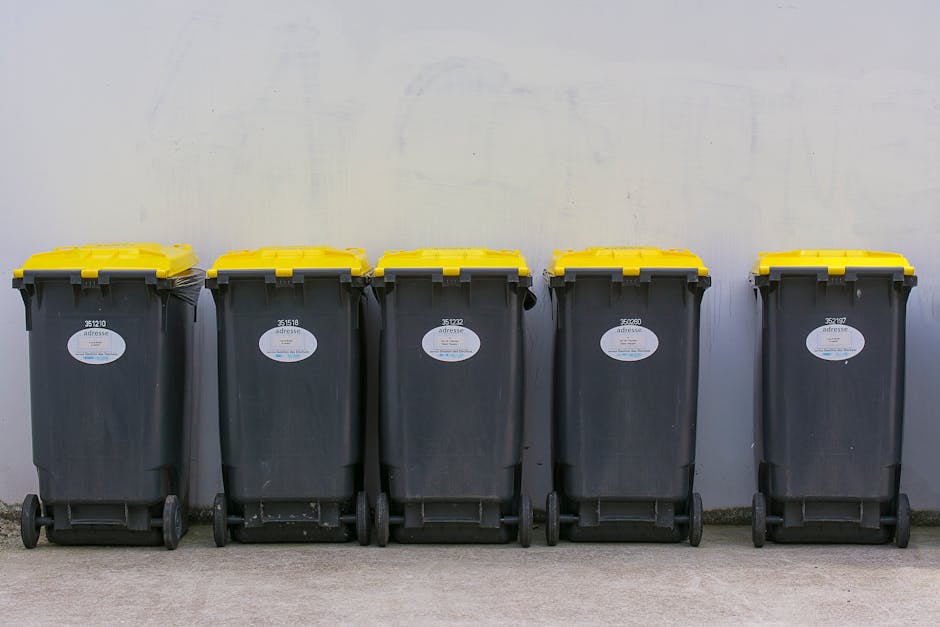 Five large wheeled rubbish bins lined up side by side against a plain, light grey wall on a concrete surface. The bins are made of black plastic with textured surfaces and are fitted with yellow lids that are closed. Each bin has a white oval label with black text and a barcode positioned on the front, indicating their designated disposal or collection categories. The bins are evenly spaced and appear to be part of an organised waste management setup, likely for a residential or commercial property in Battersea. The environment is outdoors, in what could be a driveway or service area, with minimal environmental details visible beyond the wall and concrete ground. This scene relates to private waste collection solutions offered by companies like Waste Collection Battersea, providing efficient and alternative rubbish handling for various waste types.